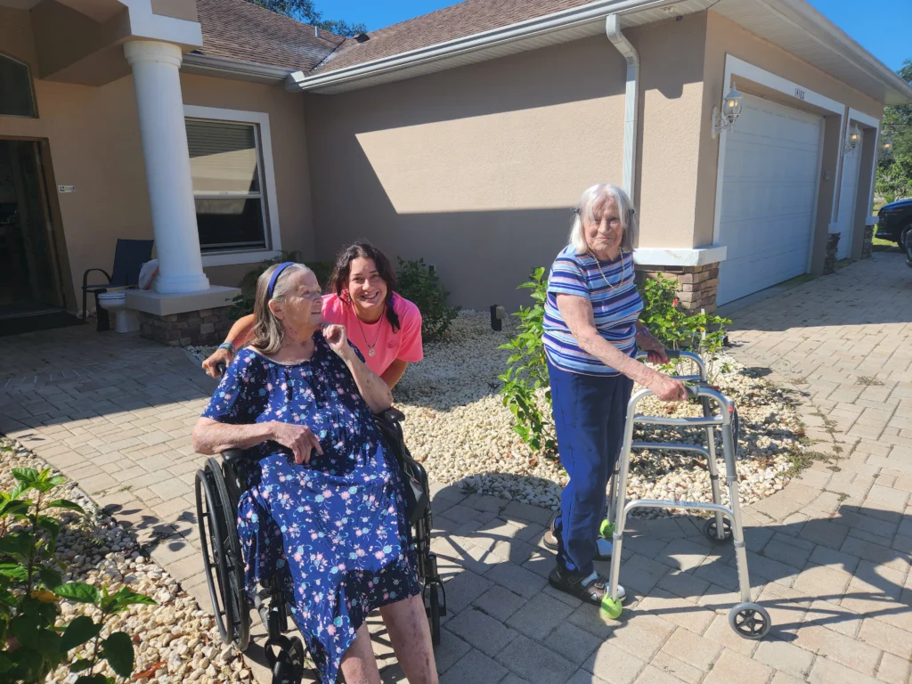 Two elderly women enjoying outdoor time at Aliviya Rose Manor, one in a wheelchair and the other using a walker, with a staff member smiling beside them in a welcoming, home-like environment.