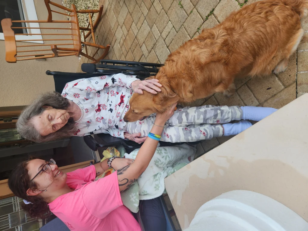 Elderly woman in wheelchair interacting with a golden retriever, while a caregiver in a pink shirt smiles and provides companionship, showcasing personalized care and emotional support at Aliviya Rose Manor.