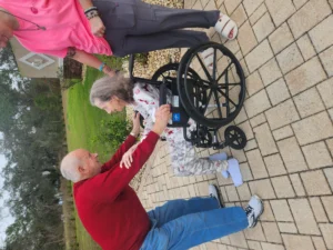 Elderly couple interacting outdoors, with a man assisting a woman in a wheelchair, emphasizing compassionate care and support for individuals with memory care needs.